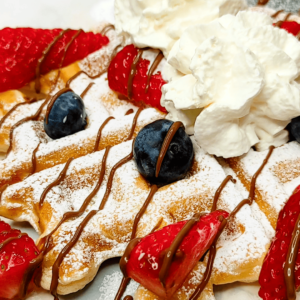 homemade waffles dusted with icing sugar and topped with fruit, melted chocolate, and whipped cream